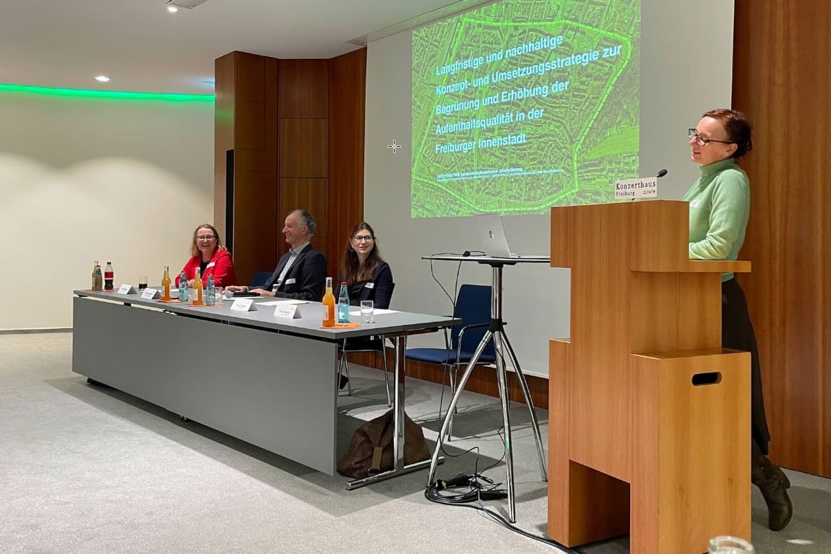 Frau an Rednerpult und drei Menschen auf dem Podium