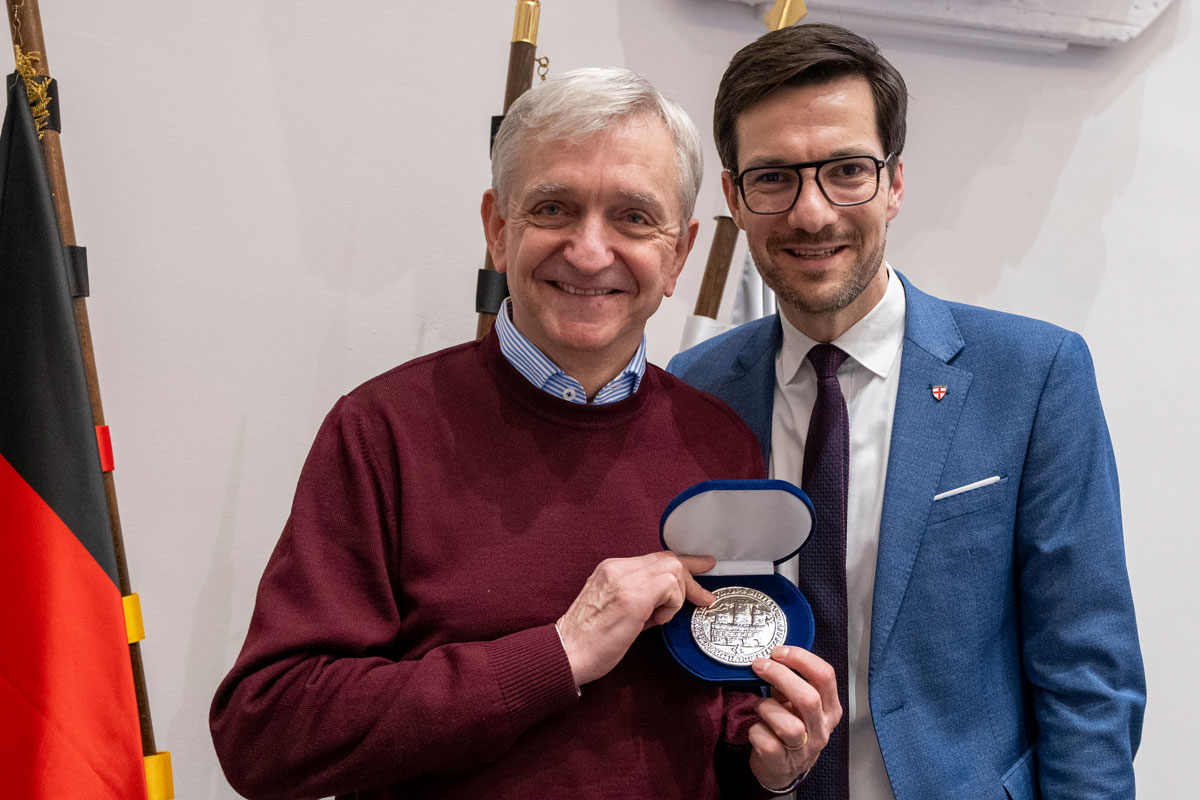 Zwei Personen grinsen in die Kamera, linke Person hält silbernes Stadtsiegel