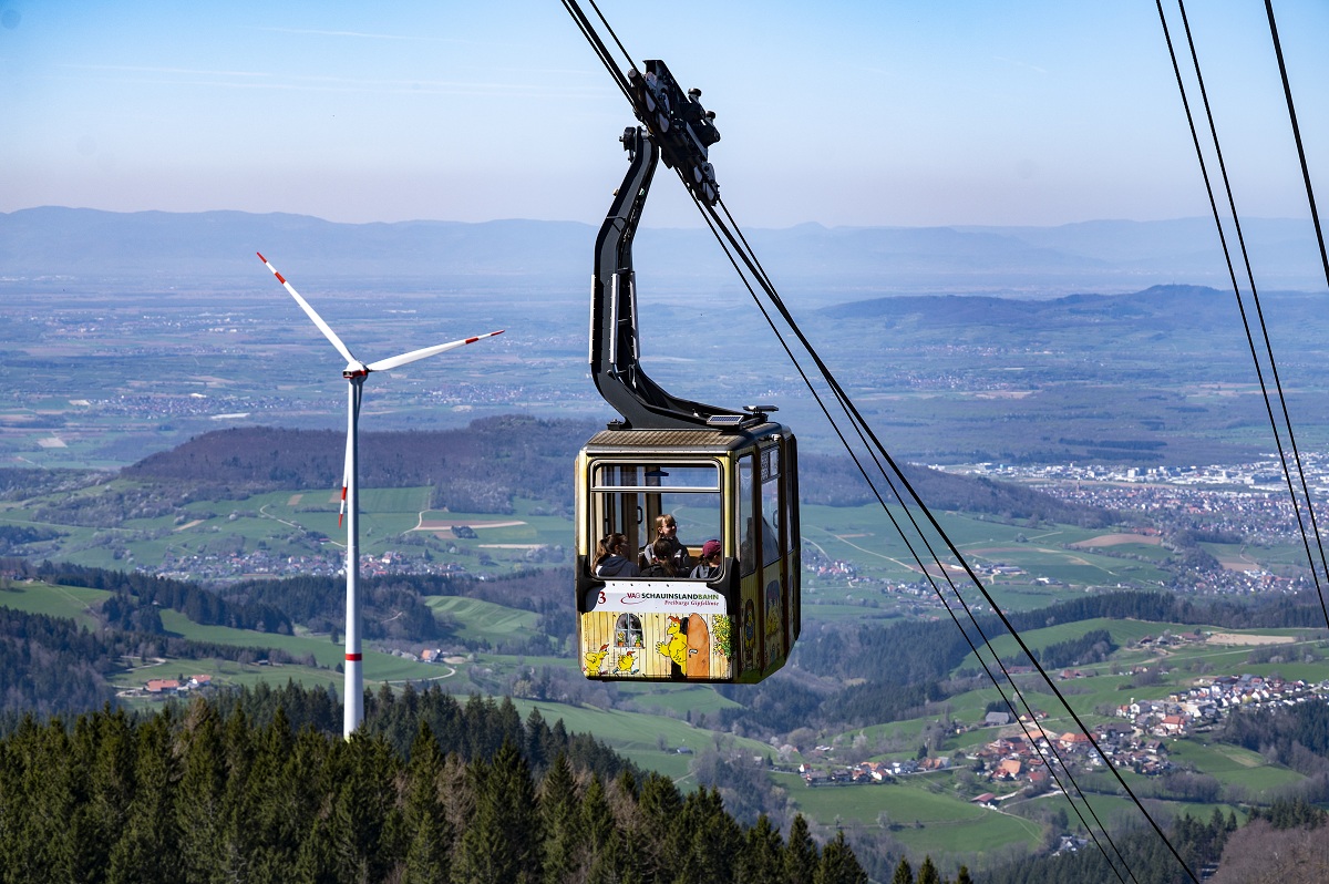 Eine Gondel der Schauinslandbahn fährt nach unten ins Tal, im Hintergrund das Panorama und ein Windrad.