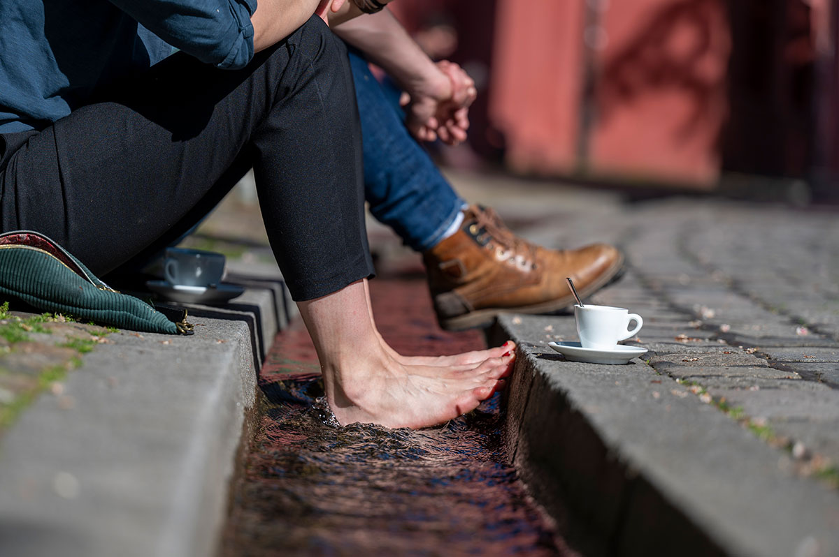 Die Beine von zwei Personen, die an einem Freiburger Bächle sitzen, eine hat die Füße im Wasser, daneben eine Kaffeetasse