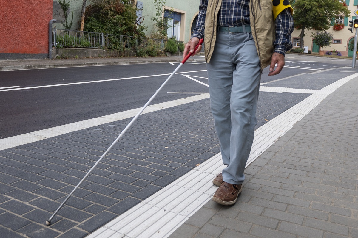 Ein blinder Mann mit Armbinde und einem Stock läuft an einer Bushaltestelle über den Leitstreifen.