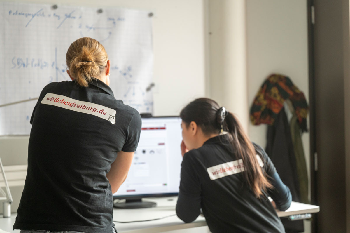 Zwei Frauen mit Aufdruck auf dem T-Shirt "Wir lieben Freiburg" schauen im Büro auf einen BIldschirm