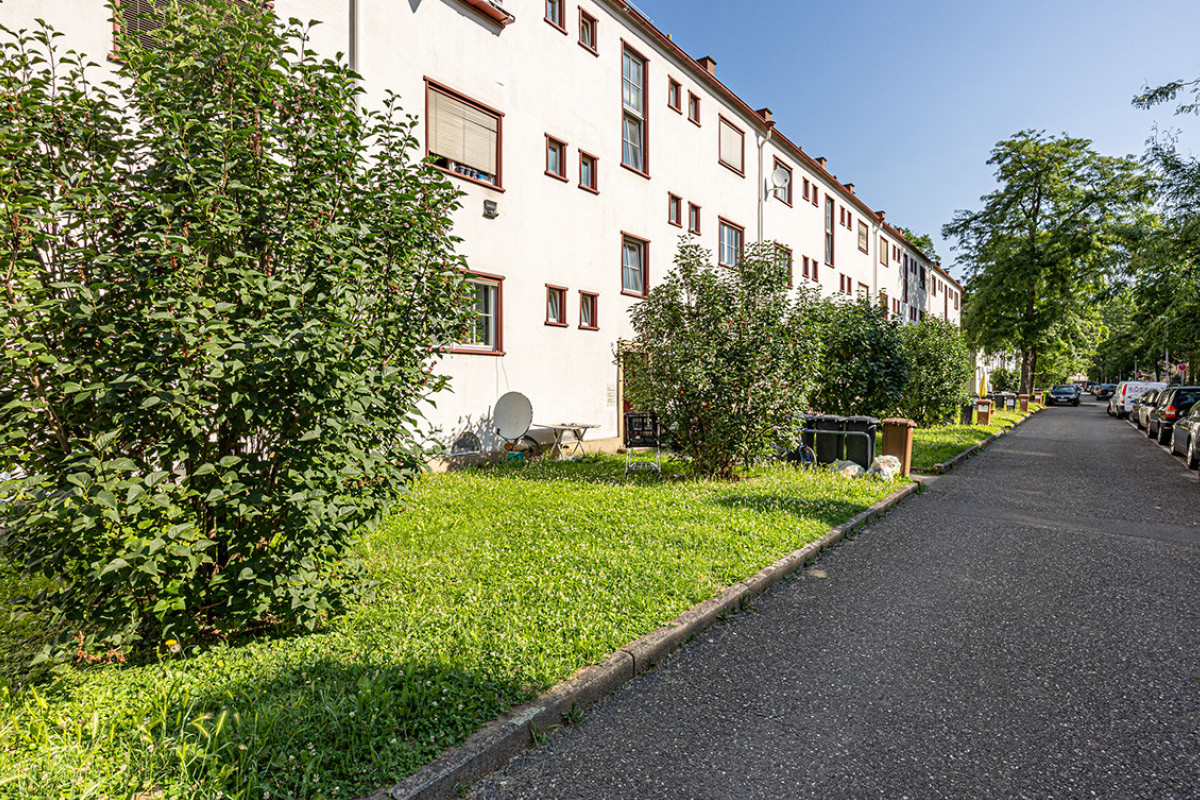 Fassade der denkmalgeschützten Gebäude zur Straße hin (Bereich 1) Alternativtext: Ein Gehweg verläuft entlang eines hell verputzten, denkmalgeschützten, dreigeschossigen Wohnhauses. Die Fenster sind rotbraun umrahmt. Zwischen Fassade und Gehweg liegt ein grüner Vorgarten mit Rasen und dichten Sträuchern. Mülltonnen stehen am Straßenrand; am Gehweg entlang sind mehrere Autos geparkt. Die Straße sieht man nicht.