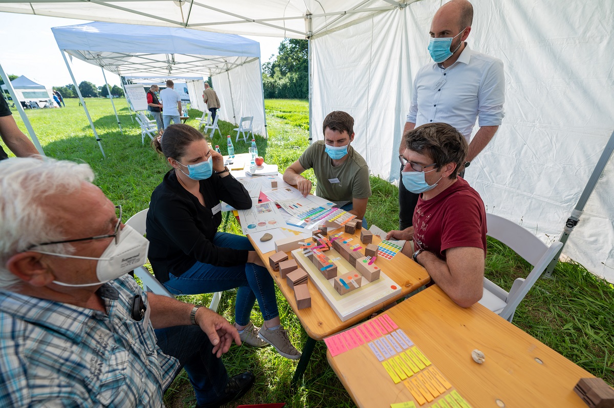 Fünf Personen mit Maske sitzen um einen Tisch mit einem Holzklötzchen-Baublockmodell