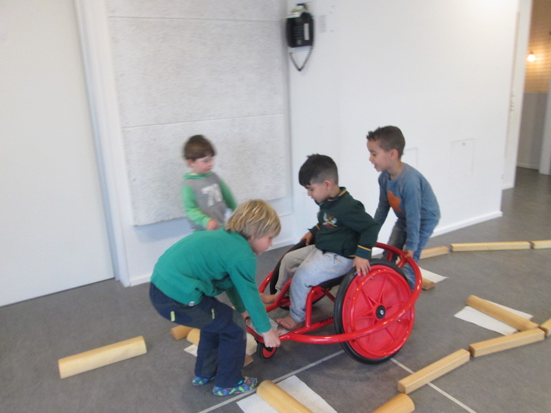 Einem Junge mit Rollstuhl wird von zwei weiteren Jungen geholfen, eine Barriere im Parcours zu überwinden,