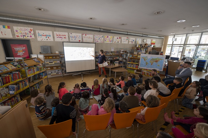 Viele Kinder sitzen auf Stühlen und Sitzkissen und schauen zu einer Leinwand.