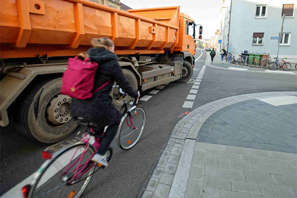 Symbolfoto: Ein LKW schneidet beim Rechtsabbiegen eine Radlerin, die sich im toten Winkel rechts neben dem Fahrzeug befindet.