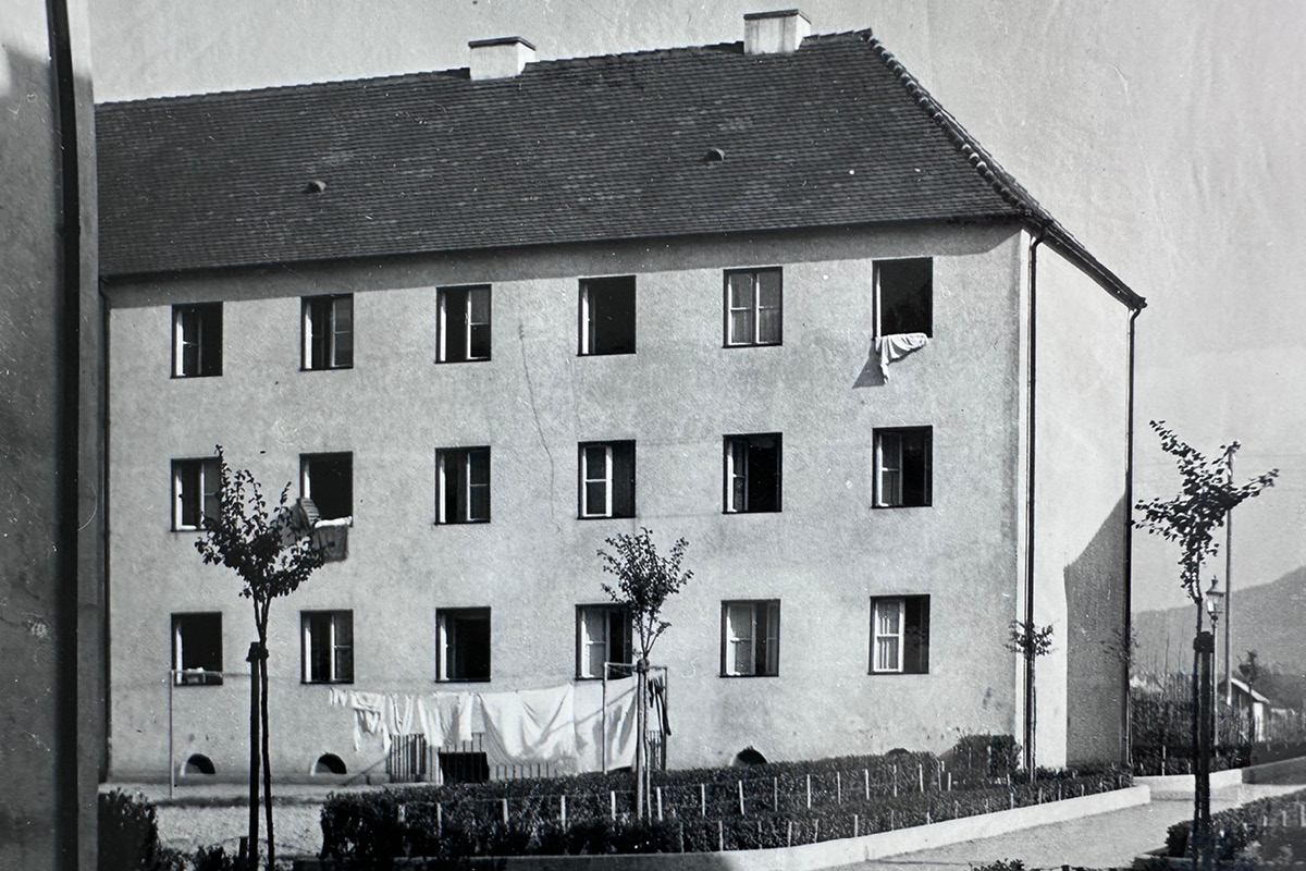 Historisches Foto des Innenhofs zwischen den denkmalgeschützten Gebäuden im Bereich 1 (ca. 1950er Jahre) Historische Schwarz-Weiß-Aufnahme des Innenhofs zwischen den denkmalgeschützten Gebäuden, etwa aus den 1950er-Jahren. Zu sehen ist ein schlichtes, dreigeschossiges Wohnhaus mit steilem Ziegeldach und gleichmäßig angeordneten Fenstern. Im schmalen Vorgarten stehen junge Bäume und niedrige Hecken; Wäsche hängt vor der Fassade zum Trocknen.