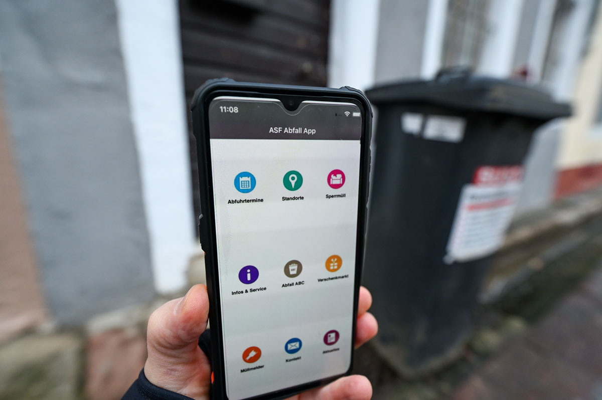 Person hält Handy ins Bild, darauf geöffnet: Abfall-App, im Hintergrund Mülltonne