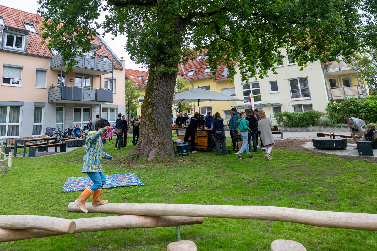 Kind balanciert im Vordergrund, im Hintergrund Bänke und Tische mit Menschen unter einem großen Baum