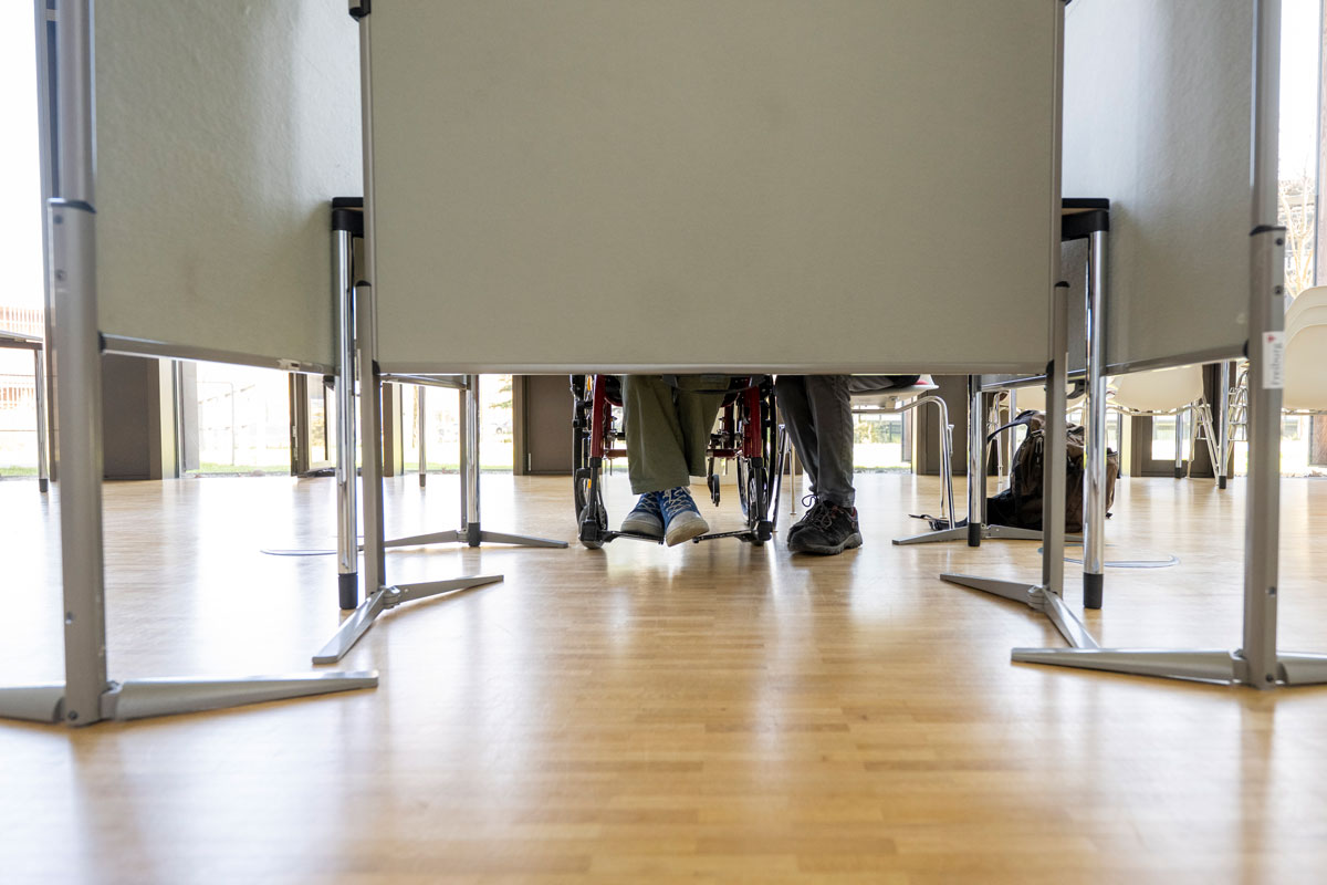 Zwei Personen sitzen in Wahlkabine - eine Person mit einem Rollstuhl