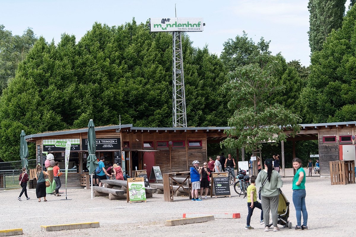 Eingansbereich des Mundenhofs, ein Sandplatz mit Holzbänken, Tischen, Kiochk und Turm mit Mundenhof-Schriftzug an der Spitze.