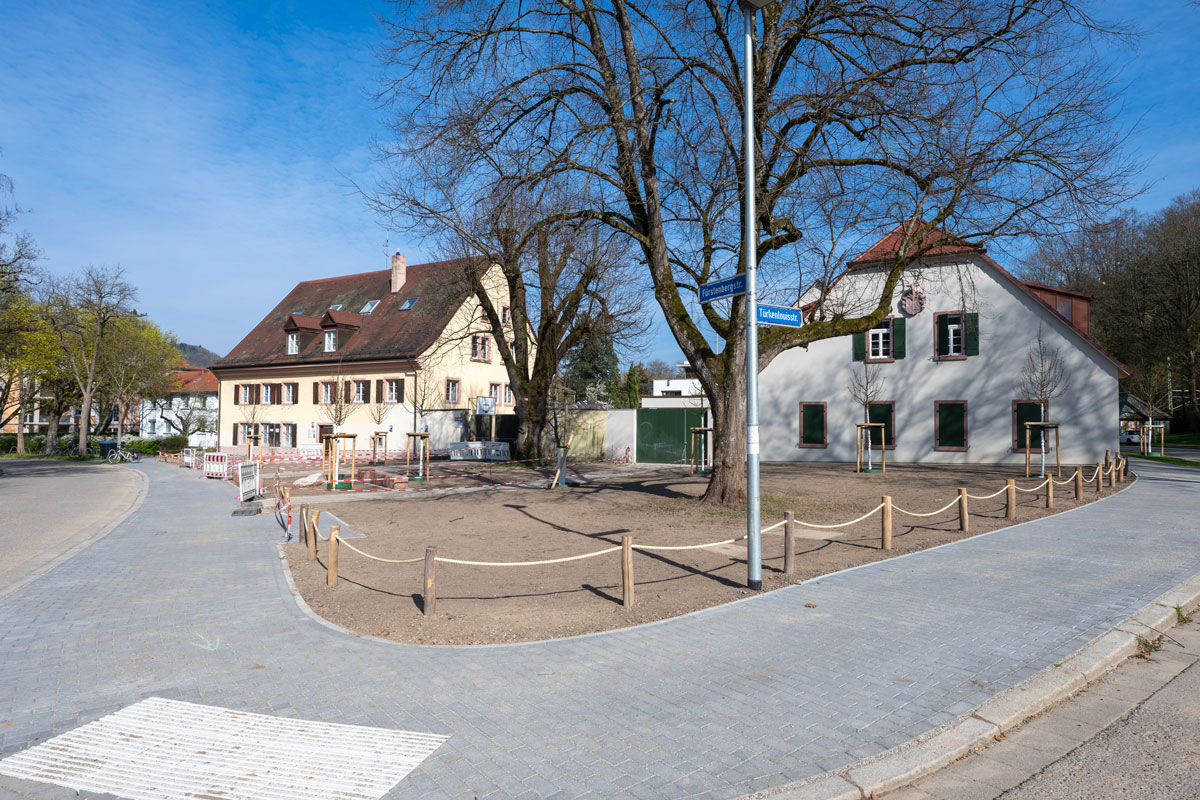 Große entsiegelte Fläche mit Bäumen, im Hintergrund Wohnhäuser