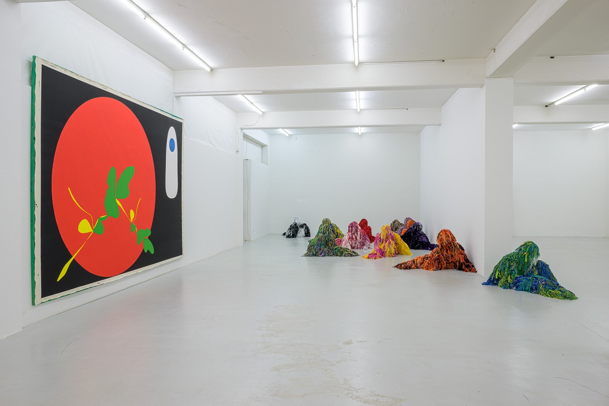 Ausstellungsansicht mit einer großen Malereie mit einem großen roten Kreis von Pia Rosa Dobrowitz und vielen kauernden Figur in bunten Tarnanzügen von Yongkuk Ko