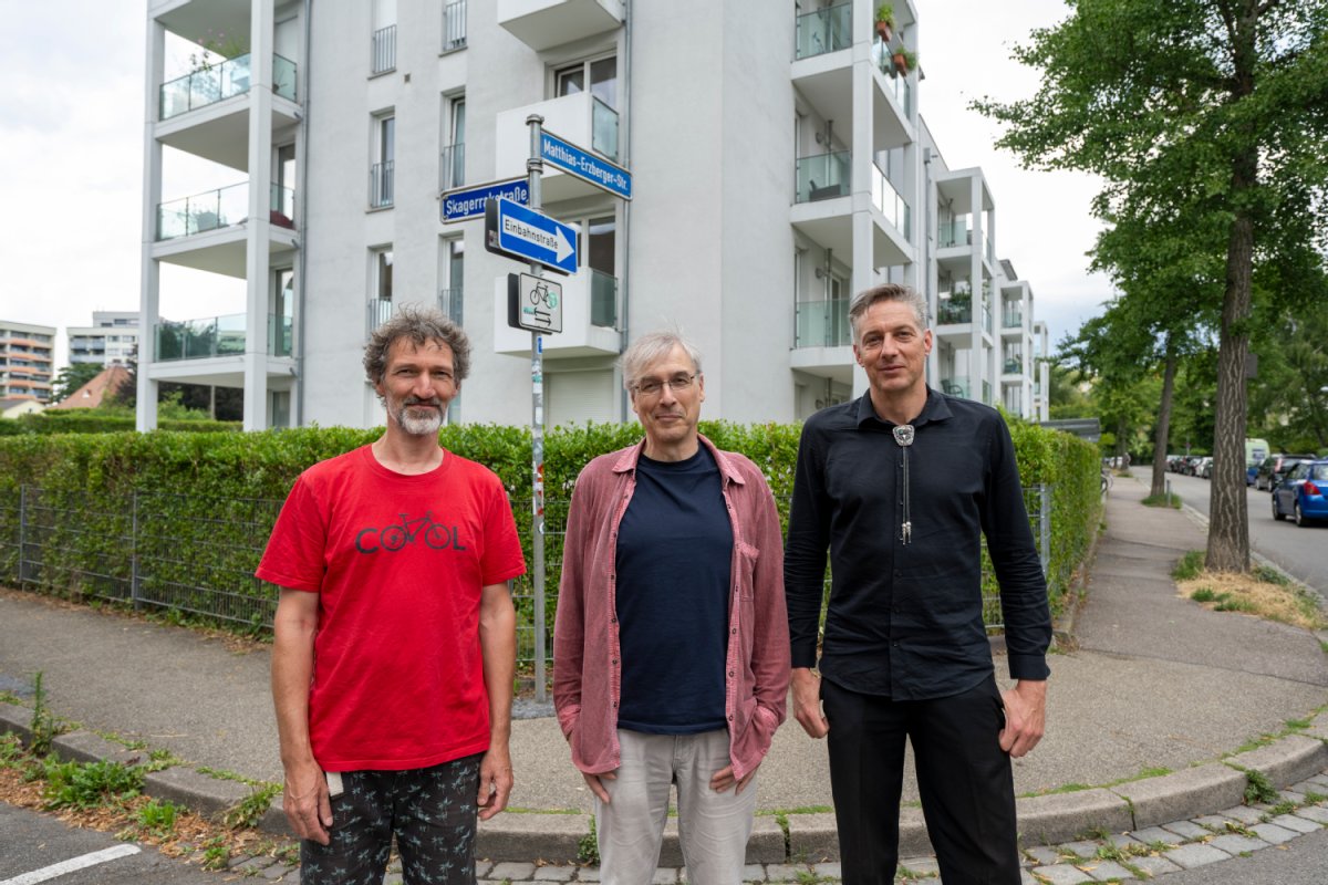 Drei Männer stehen vor einem Wohngebäude an einer Kurve.