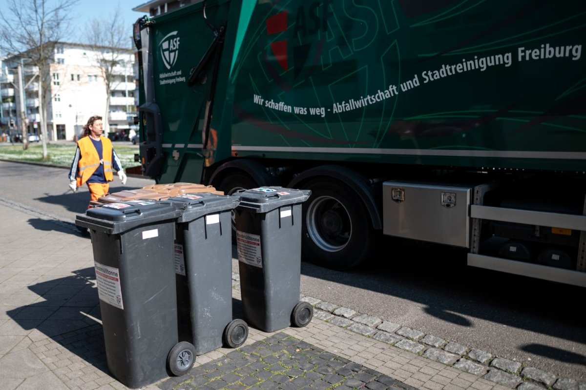 Restmüll- und Biotonnen auf der Straße, dahinter ein LKW der ASF. Daneben ein Müllwerker.