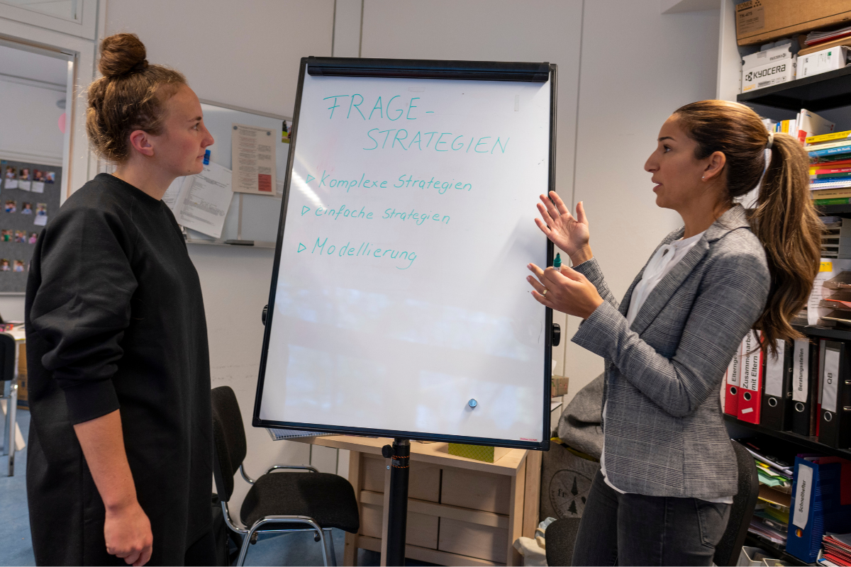 Eine Referentin bespricht mit einer Fachkraft Fragestrategien vor einem Whiteboard.