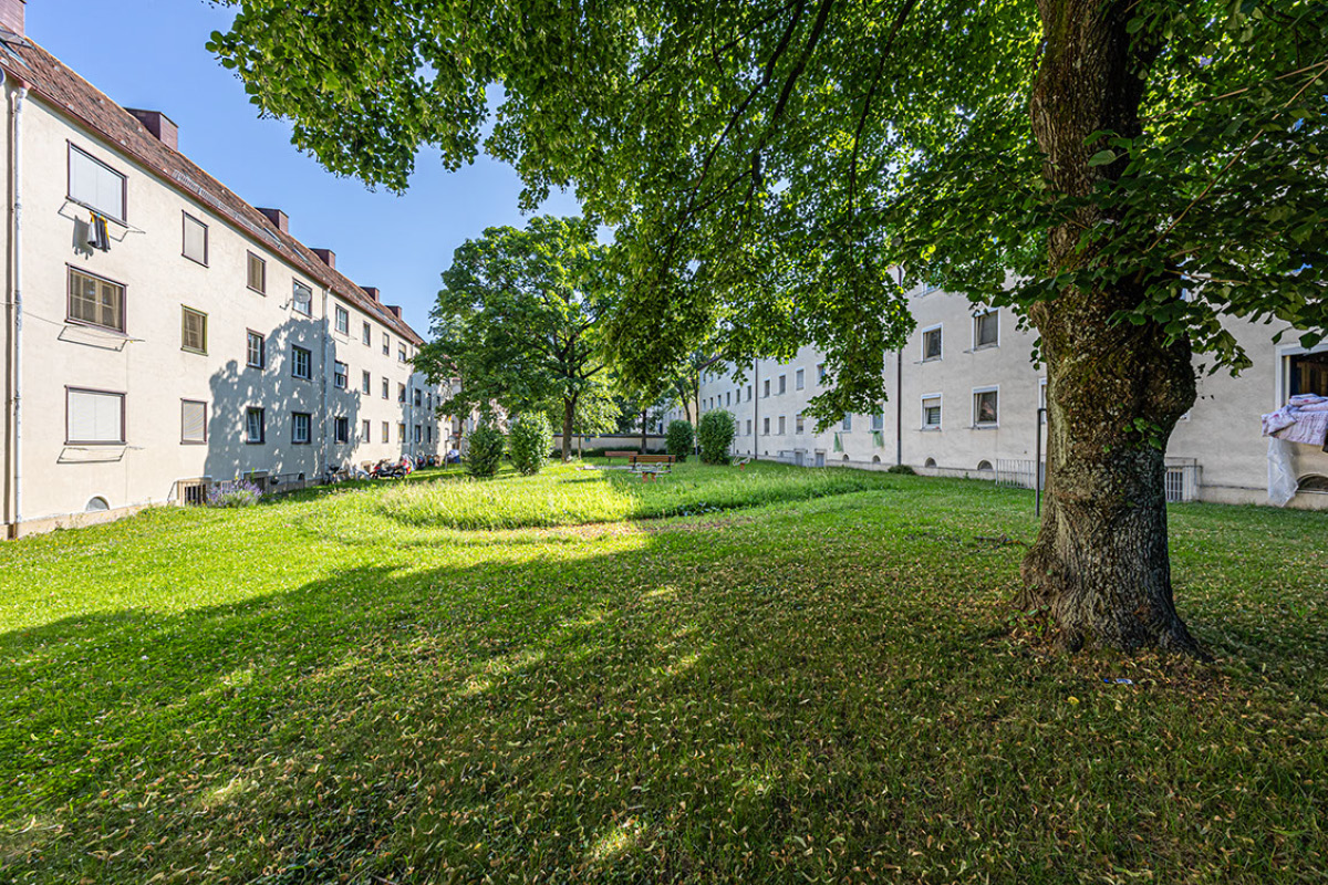 Grüner Innenhof zwischen den denkmalgeschützten Gebäuden (Bereich 1) Blick in einen begrünten Innenhof, den helle dreigeschossige und denkmalgeschützte Wohnhäuser rahmen. Große alte Bäume spenden Schatten; in der Rasenfläche steht eine Bank.