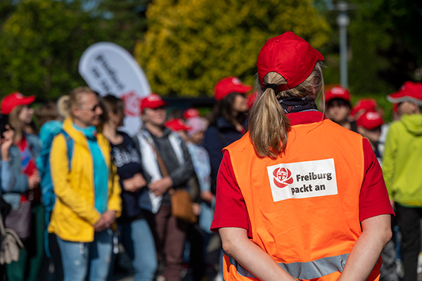 Frau mit orangener Warnweste mit der Aufschrift "Freiburg packt an", im Hintergrund viele Menschen