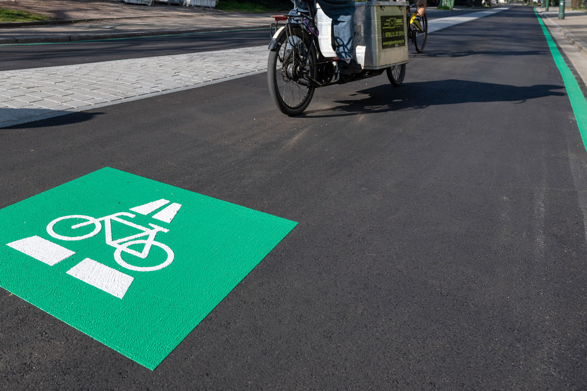 Großes, grünes Radpiktogramm auf der Straße, im Hintergrund Fahrradfahrende