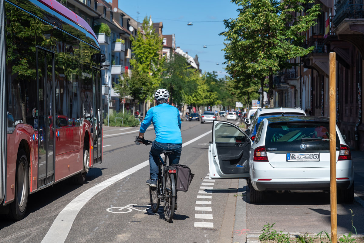 Radfahrer auf einem Radweg zwischen Bus und parkendem Auto mit geöffneter Tür.