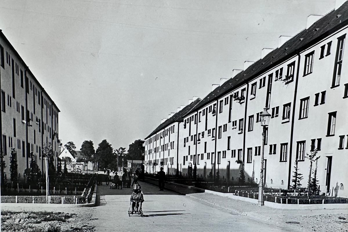 Historisches Foto des Straßenraums zwischen den denkmalgeschützten Gebäuden im Bereich 1 (ca. 1950er Jahre) Historische Schwarz-Weiß-Aufnahme des Straßenraums zwischen den denkmalgeschützten Gebäuden, etwa aus den 1950er-Jahren. Eine breite Straße führt zwischen langen, dreigeschossigen Wohnhäusern hindurch. Vorgärten mit niedrigen Einfassungen säumen die Häuser; mehrere Menschen sind auf der Straße und den Wegen unterwegs. Im Vordergrund sitzt ein Kind auf einem Stuhl mit Rädern, rechts steht eine historische Straßenlaterne.
