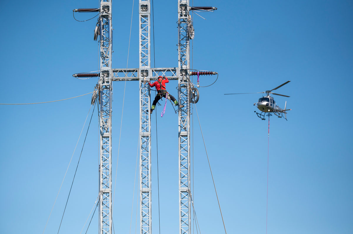 Eine Person bringt Kabel auf einem Strommast an, im Hintergrund ein Helikopter