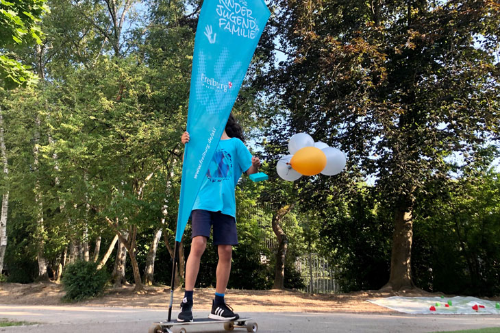 Jugendlicher fährt auf einem Longboard / Skateboard und hat Luftballone sowie eine große Beachflag (Beschriftung: Amt für Kinder, Jugend und Familie und www.freiburg.de/aki) in der Hand. 