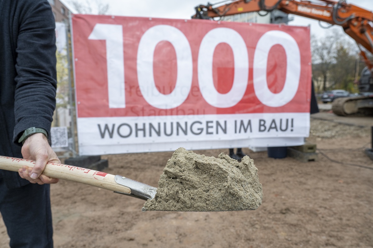 Eine Schaufel mit Sand bei einem Spatenstich, im Hintergrund ein Banner mit Aufschrift: "1000 Wohnungen im Bau!"